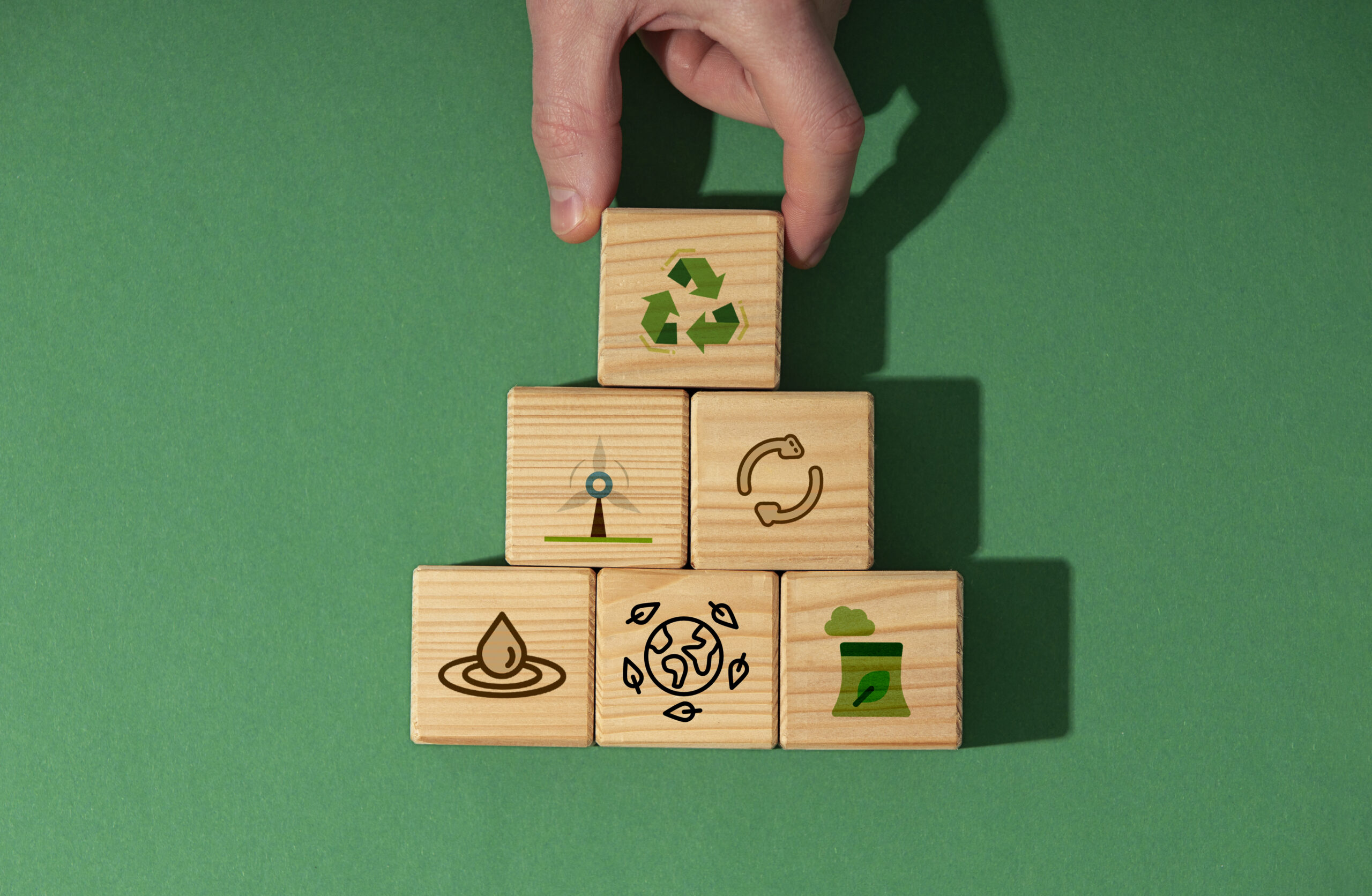 Wooden building blocks on a green background. Lying flat, face up to the camera, stacked on top of each other and tiered. Each block has a different illustrated symbol representing an aspect of sustainability.