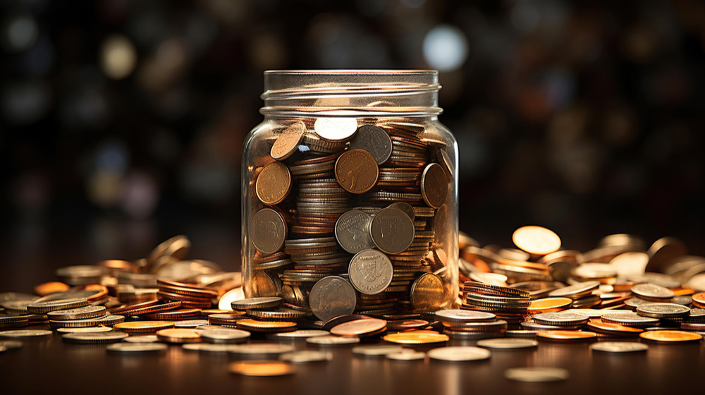 A clear jar full and surrounded by coper pennies sits on a dark wood table. The background is dark and out of focus.