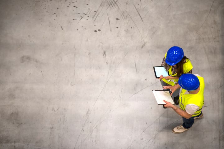 Aerial shot of two workers, one male, one female. Both in yellow high vis jackets and blue hard hats. Standing to the right of the shot, the rest of the space is empty concrete floor. The male figure is holding a paper and a pen, the female figure is holding a tablet.